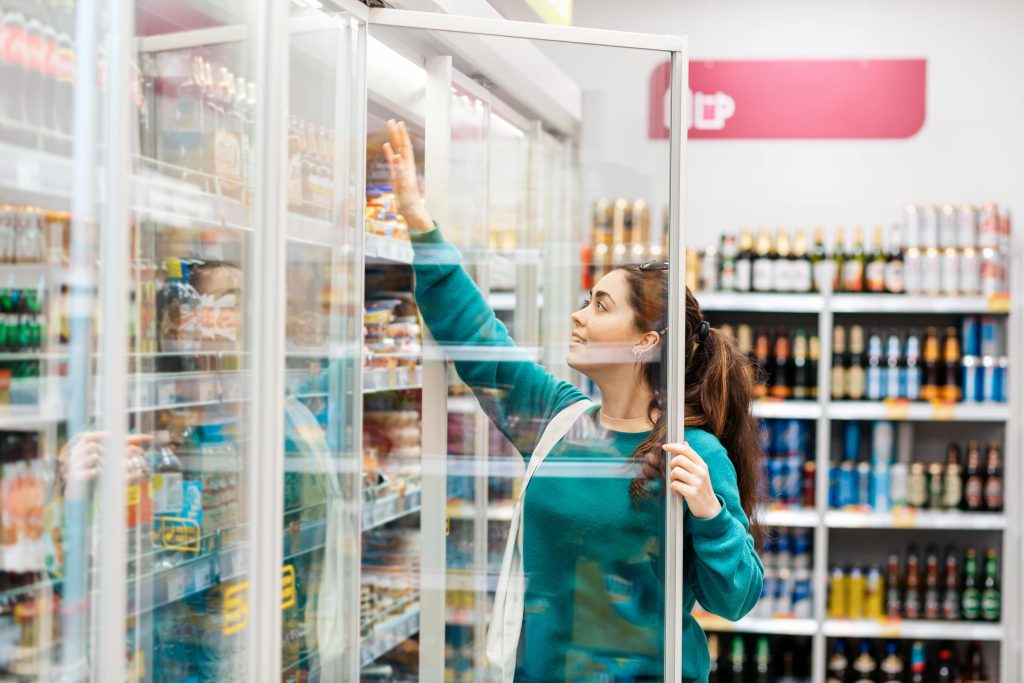 A customer pulling a product off the top shelf of a grocery store cooler